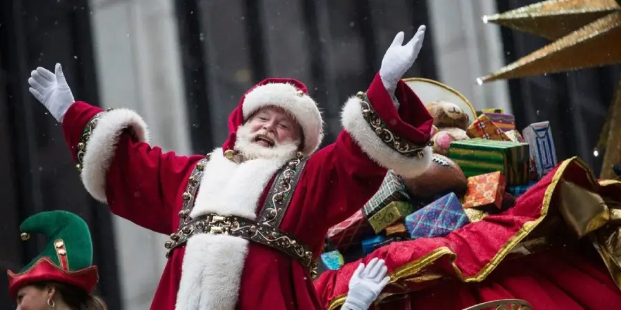 Santa Claus, dressed in a red and white suit, joyfully raises his arms while standing in front of a sleigh filled with colorful wrapped presents during a festive outdoor parade.