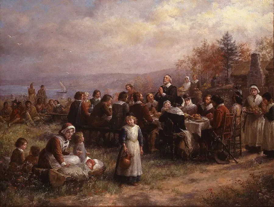 A historical painting depicting Pilgrims and Native Americans gathered outdoors, sharing a meal at long tables with food, set in an autumn landscape near water.