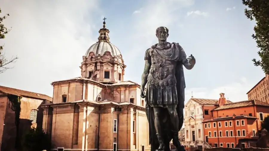 A bronze statue of a Roman figure stands before a historic domed church and other old buildings in Rome, Italy, under a partly cloudy sky.