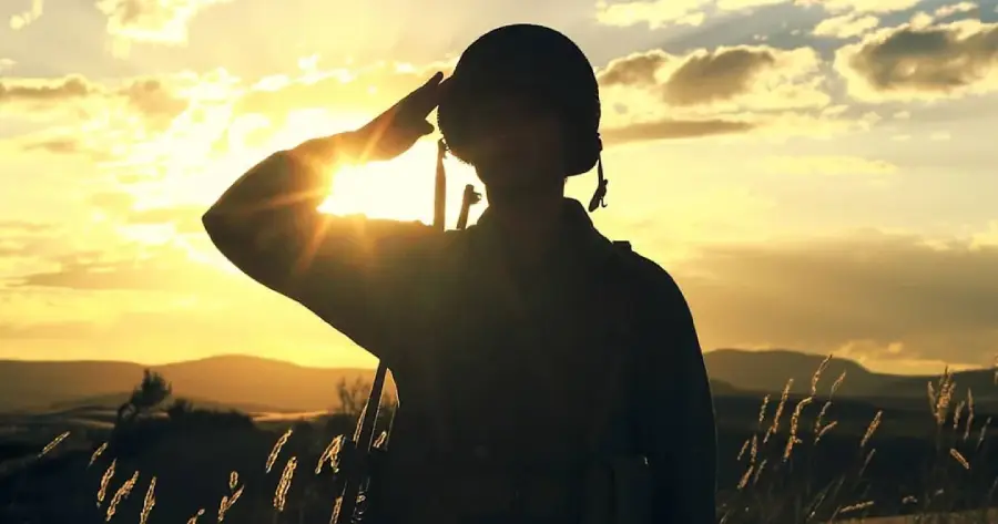 A soldier in uniform salutes at sunset, standing in a field with tall grass and hills in the background, the sun shining behind them.