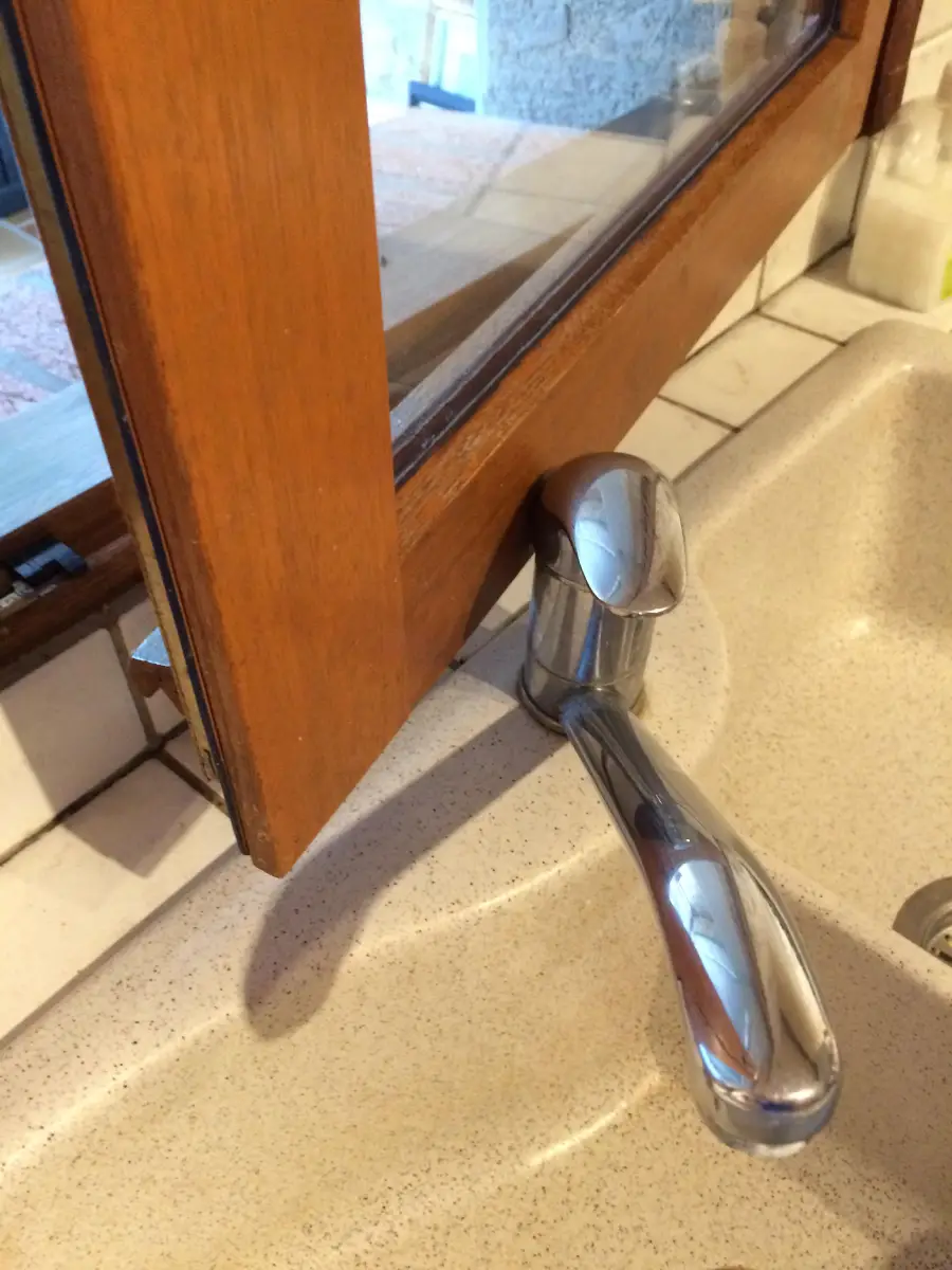 A kitchen faucet is installed too close to a low window, making it impossible to open the window fully without hitting the faucet handle.
