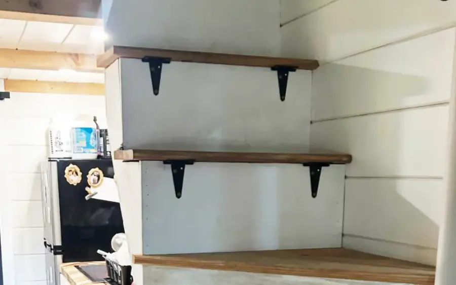 Wooden stairs with white risers, black metal brackets, and brown treads in a small, white-paneled kitchen area. A black refrigerator is visible in the background.