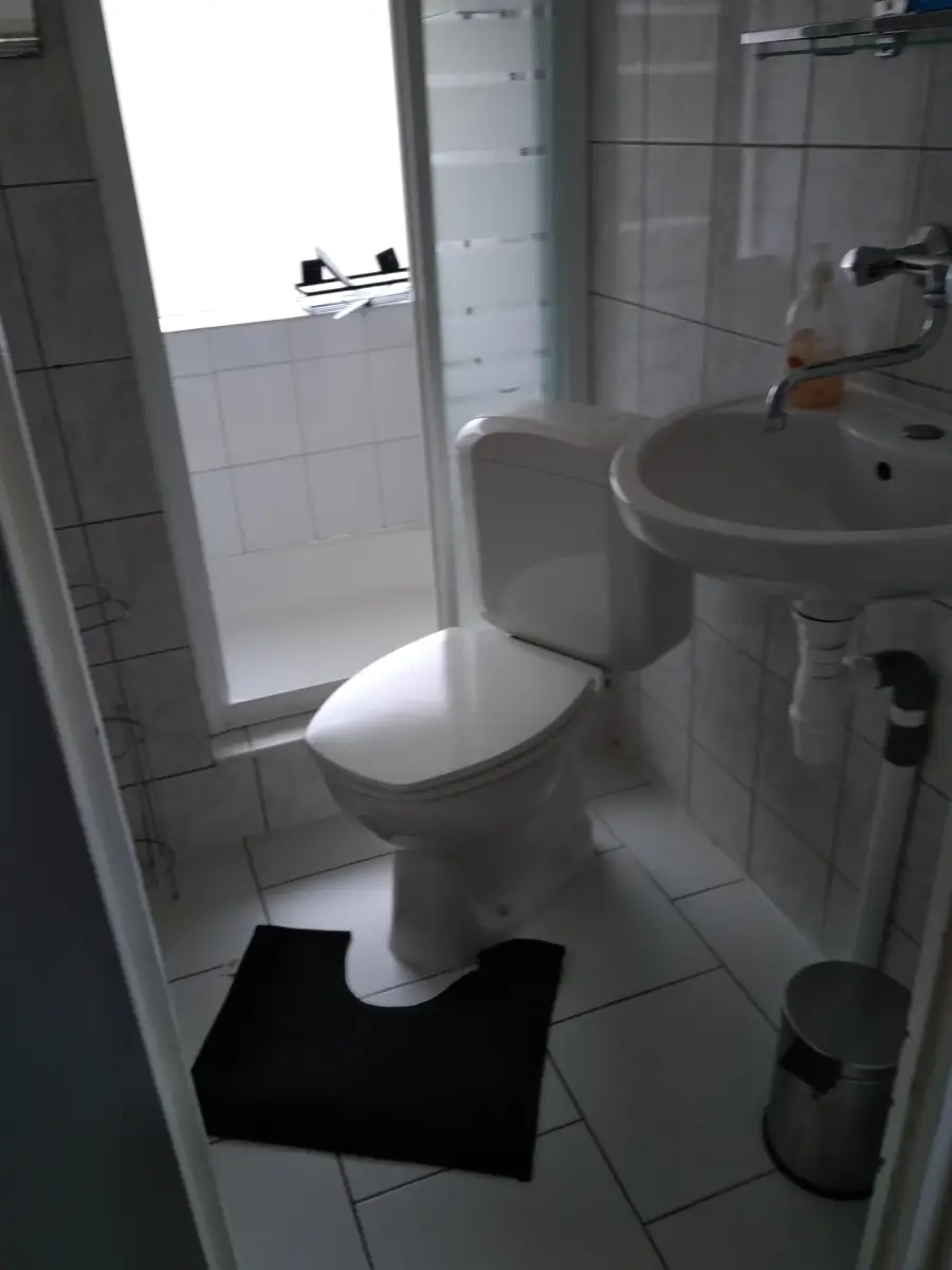 A small bathroom with a toilet, wall-mounted sink, and a shower. There is a black floor mat in front of the toilet and a silver trash bin beside it. The walls and floor are tiled in white.