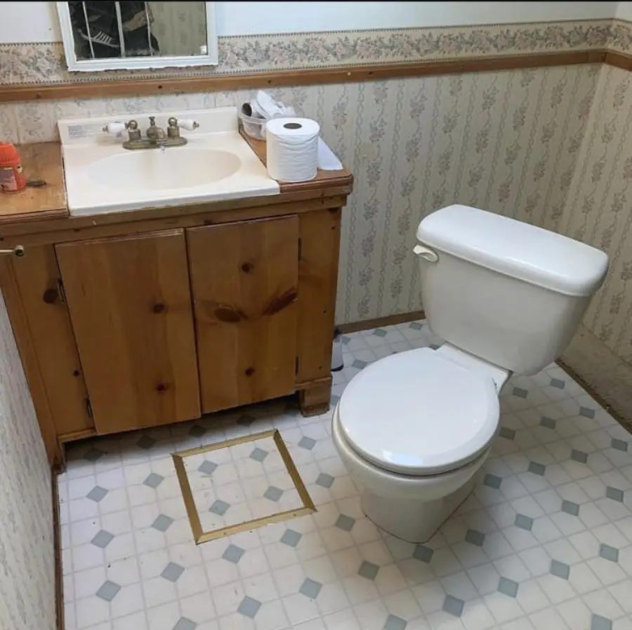 A bathroom with a white toilet next to a wooden sink cabinet. The floor has tiled linoleum with a square outline, possibly indicating where something used to be. Toilet paper and soap are on the sink.