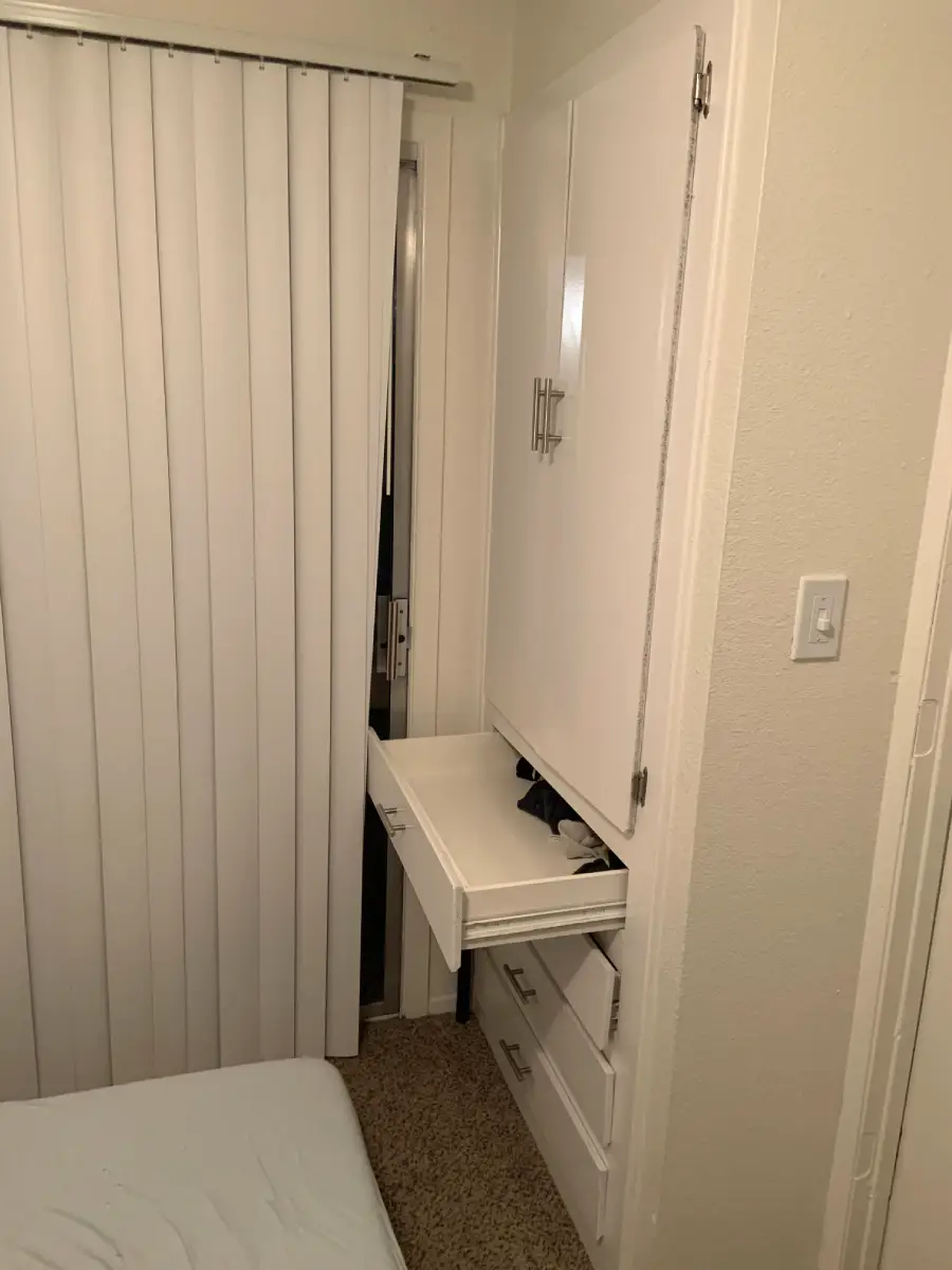 A small bedroom with a built-in dresser and open drawer blocking the sliding door to a balcony. The bed is very close to the dresser and sliding door, showing tight space.