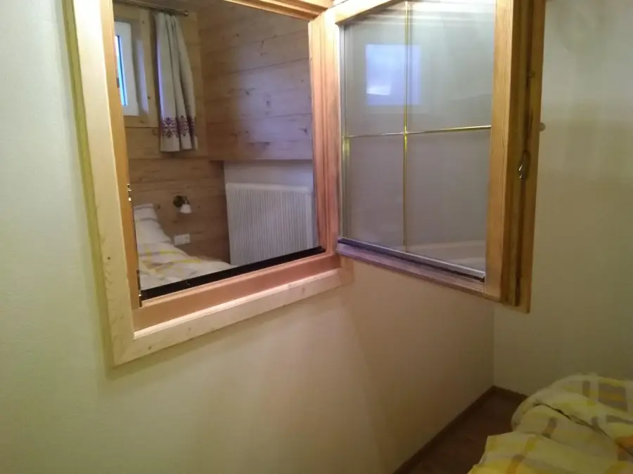 A wooden-framed indoor window opens from one room into another, revealing a bed with yellow plaid sheets, a radiator, and wood-paneled walls with a small external window and floral curtains.