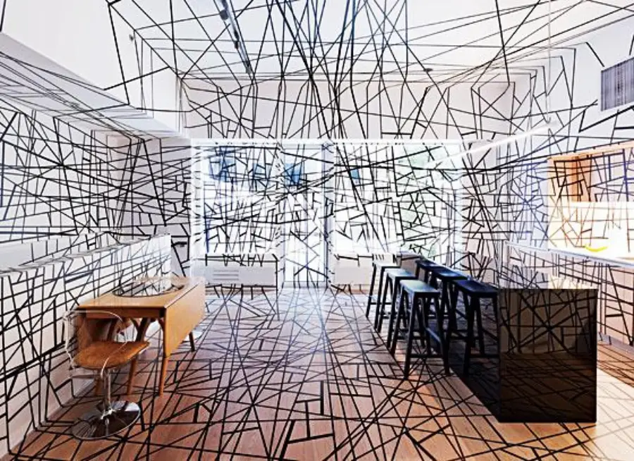 A modern room features black crisscrossing lines across the floor, walls, ceiling, and furniture. The space includes a desk with a chair, a bar with stools, and white shelves, giving it an abstract, artistic appearance.