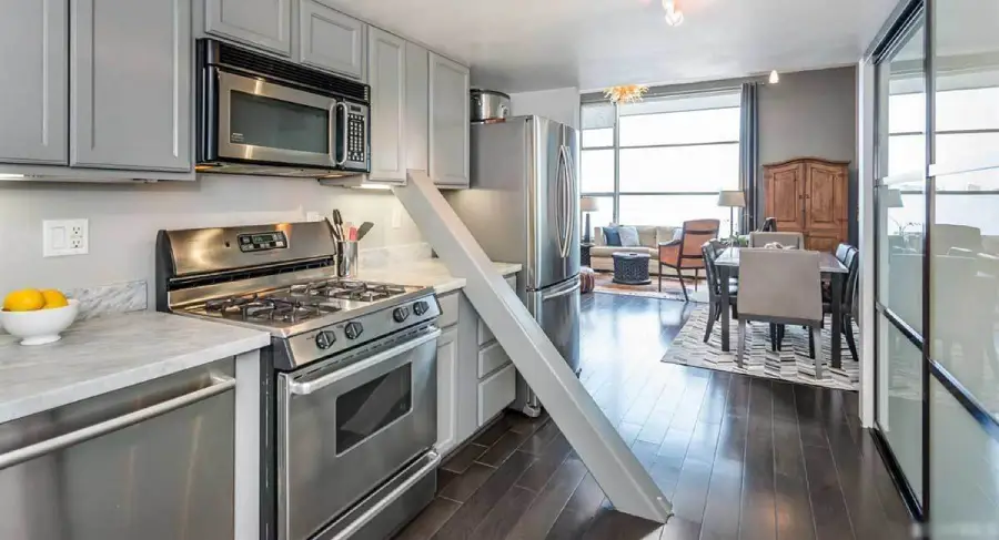 Modern kitchen with stainless steel appliances and gray cabinets, featuring a white marble countertop. A tall cabinet door is leaning open, revealing a dining and living area in the background.