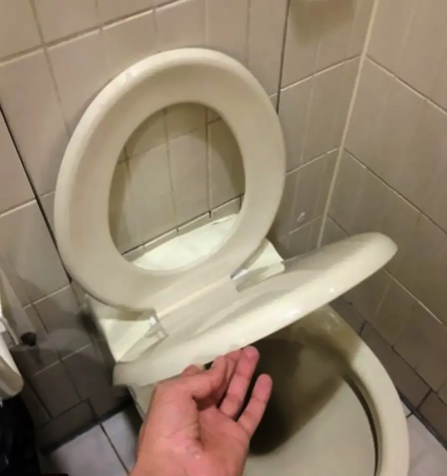 A hand lifts a toilet seat, revealing another identical seat directly beneath it, both in a beige-tiled bathroom.