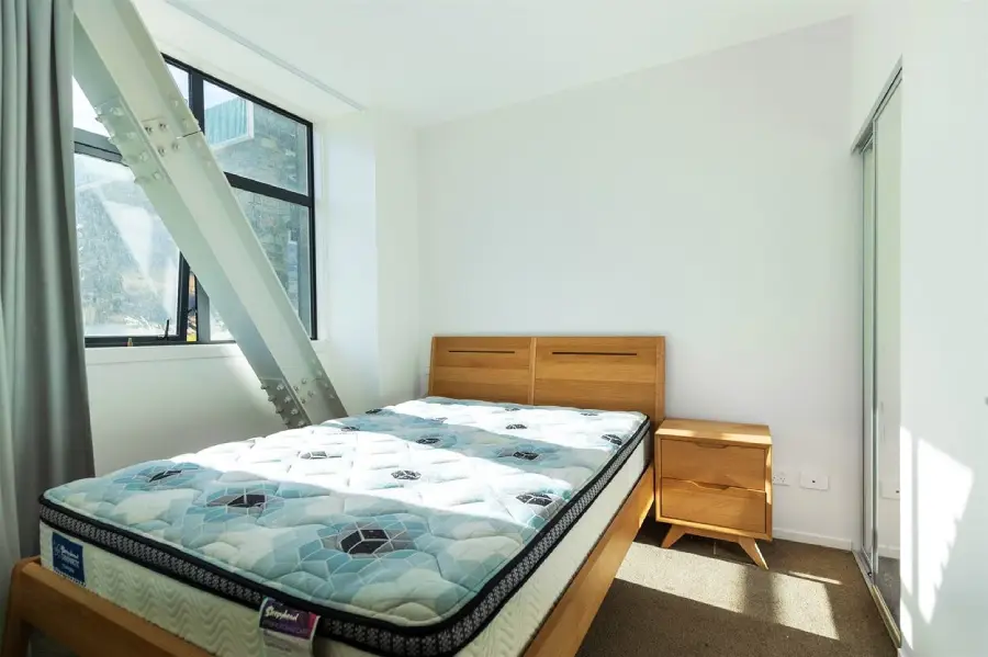 A bright bedroom with a wooden bed frame, mattress, and matching nightstand. Large window with gray curtains, exposed metal beam, white walls, and a closet with sliding mirrored doors. Sunlight streams into the room.