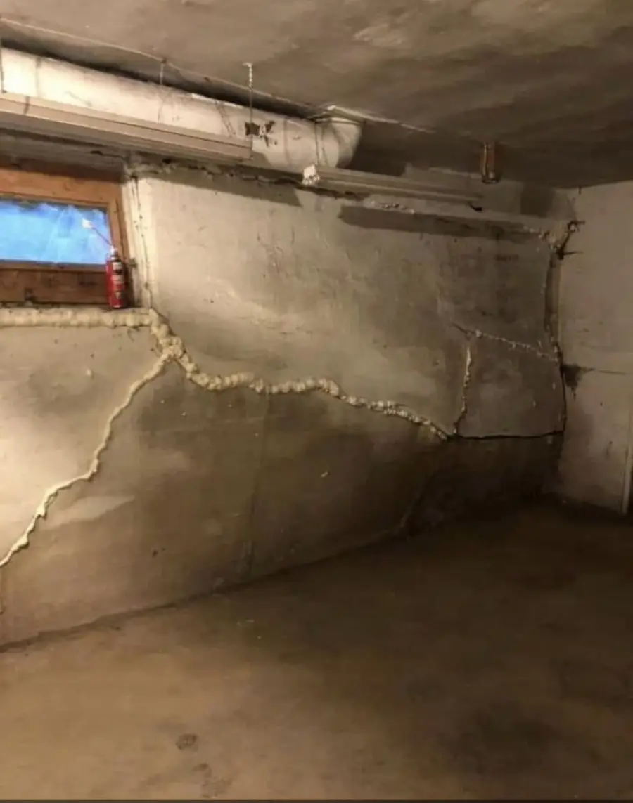 A cracked basement wall with foam sealant filling the cracks, a small window with a red canister on the ledge, and exposed pipes above. The concrete floor is bare and the room appears unfinished.