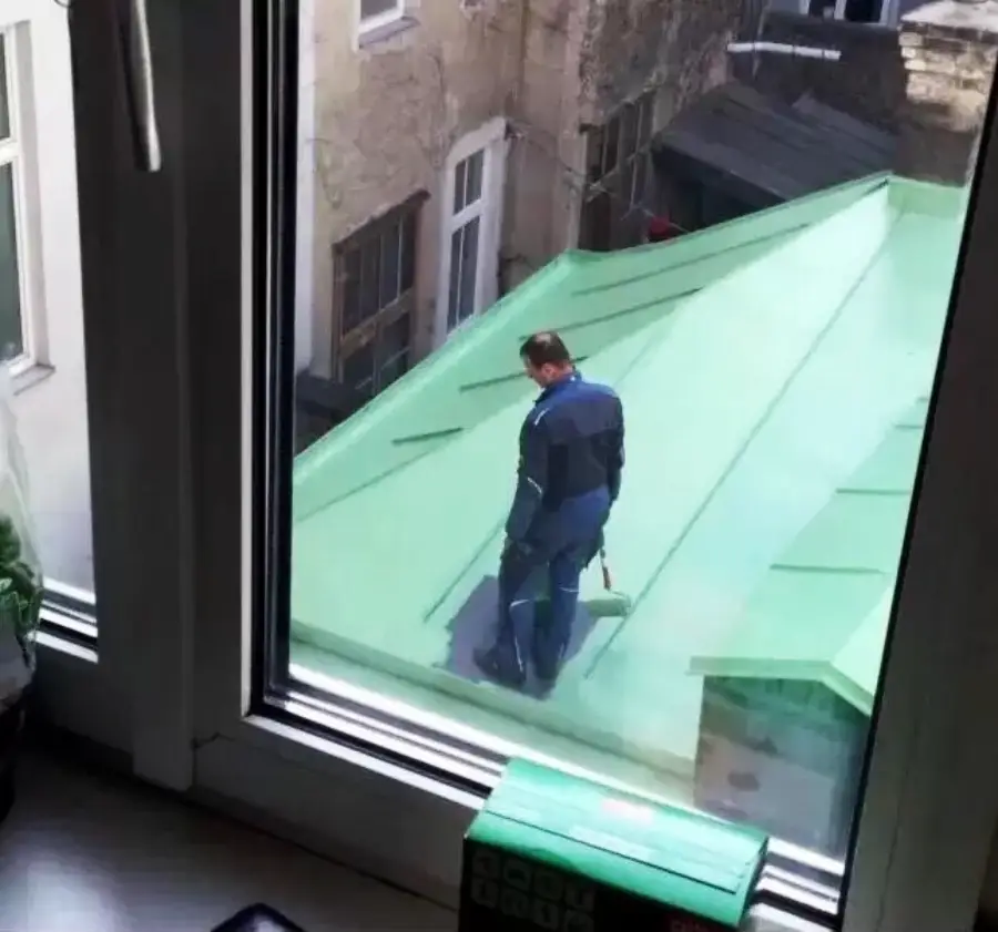 A man in blue work clothes appears to be painting or repairing a green metal roof, as seen through a window from inside a building.