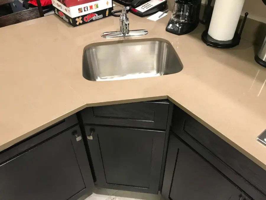 A stainless steel sink set into a tan, angled kitchen countertop with dark cabinets below. Nearby are a coffee maker, paper towels, a phone, and a board game box.