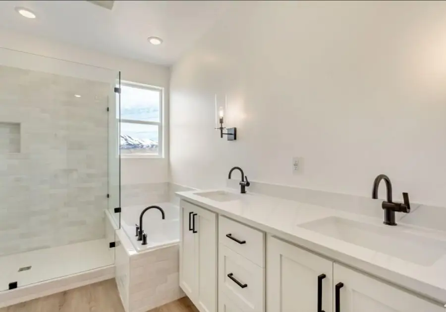 Modern bathroom with double sinks, white cabinets, black fixtures, a glass-enclosed shower, and a bathtub beside a window. Light wood floor and white walls create a clean, bright space.