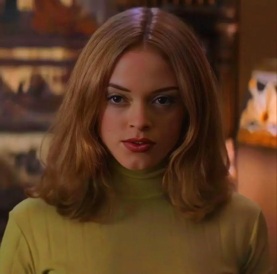 A woman with shoulder-length light brown hair and red lipstick wears a light green turtleneck. She looks directly at the camera, with a blurred, warmly lit background behind her.