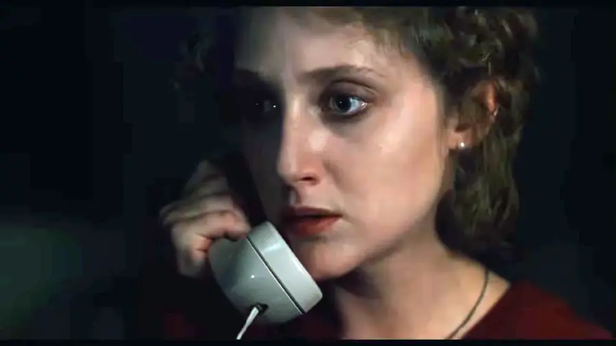 A woman with curly hair holds a white landline phone to her ear, looking anxious and tense in dim lighting, suggesting she is receiving unsettling news or is in a suspenseful situation.