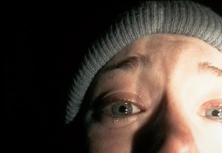 A close-up of a person's frightened face, lit from below in the dark. They wear a gray knit beanie and their eyes are wide with visible tears, conveying fear or distress.