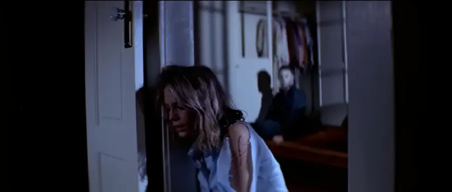 A frightened woman leans against a doorframe in a dimly lit room, while a masked figure sits menacingly in the background on a bed, partially in shadow.