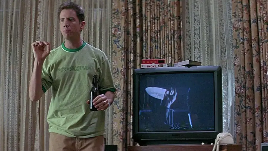 A man in a green t-shirt holding a beer stands next to a TV showing a suspenseful scene with a large knife. Several VHS tapes are stacked on top of the TV, and patterned curtains hang in the background.