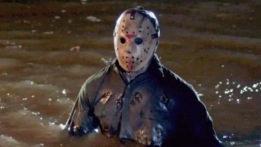 A person wearing a hockey mask and tattered, dark clothing stands waist-deep in murky water at night, creating a creepy and ominous atmosphere.