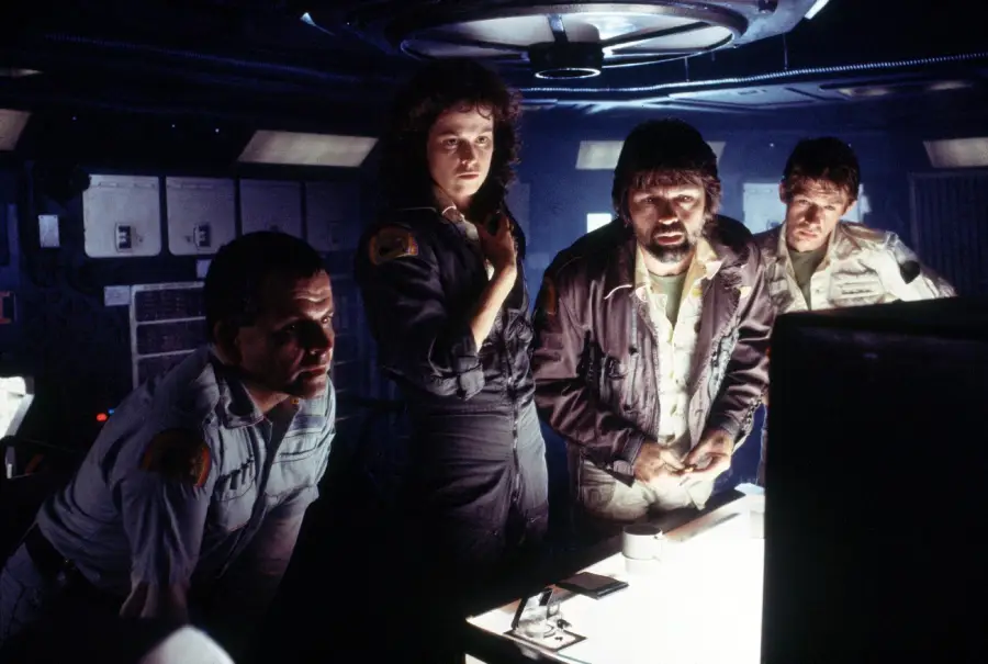 Four people in spaceship uniforms stand closely together in a dimly lit control room, looking intently at a glowing screen. The ambient lighting creates dramatic shadows on their faces.