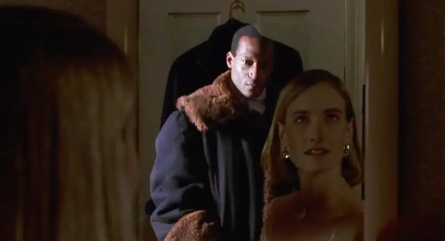 A woman looks into a mirror, her reflection visible, while a man in a dark coat with a fur collar stands behind her, appearing ominous.
