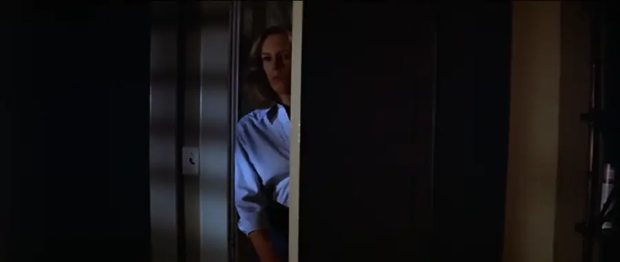 A woman in a blue shirt stands partially hidden behind a door, with only half of her face and body visible in the dimly lit scene.