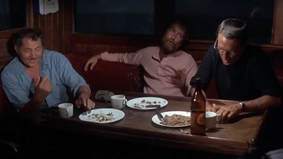 Three men sit around a wooden table in a dimly lit room, with empty plates, cups, and a brown bottle in front of them. They appear relaxed, with one man laughing and another leaning back.