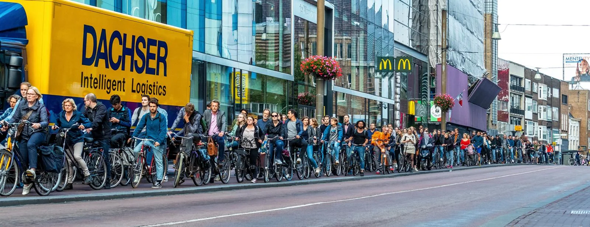 A long line of people waits with their bicycles on a city street next to a DACHSER delivery truck and storefronts, including a McDonald's, under cloudy skies.