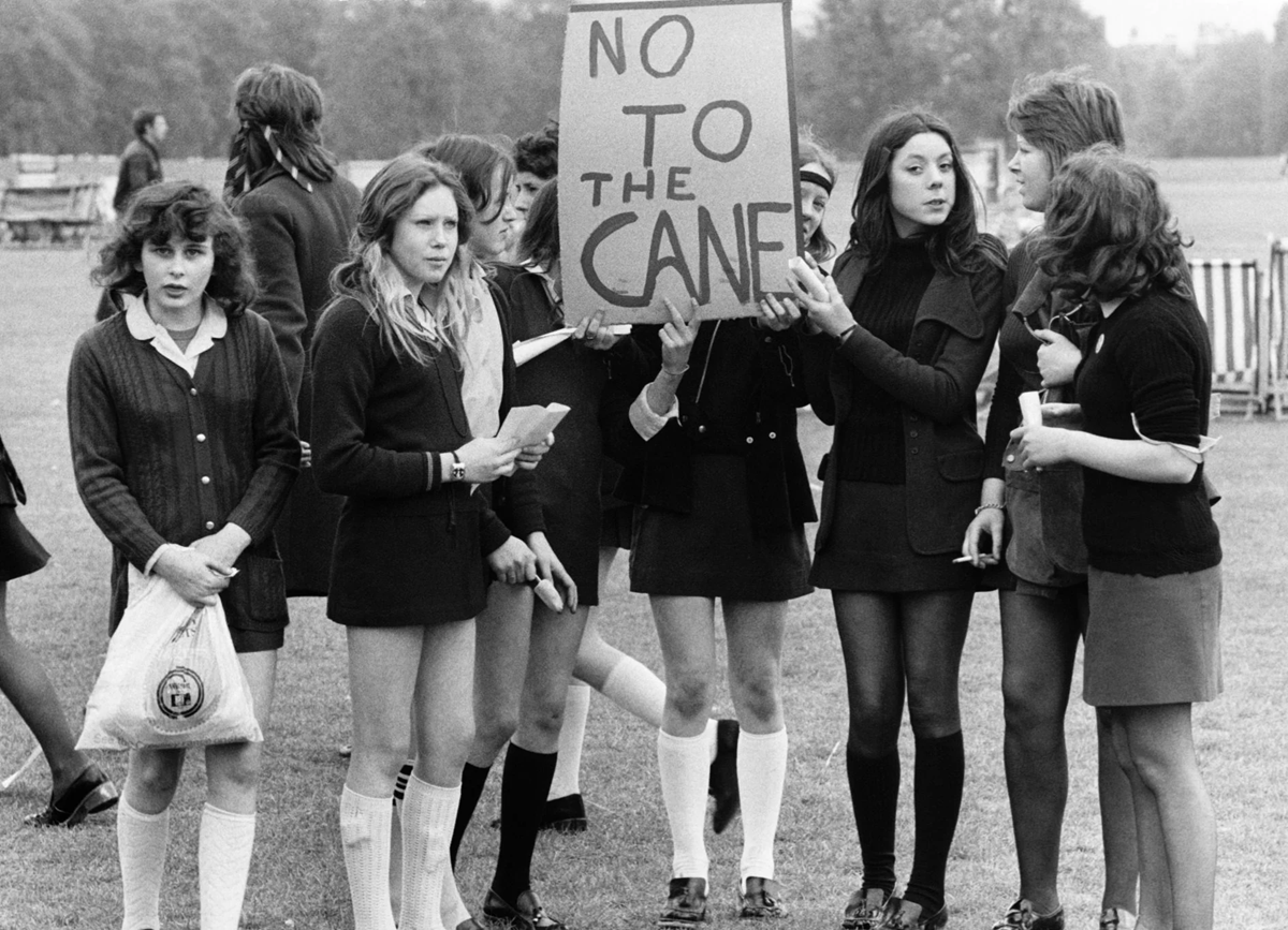 A group of schoolgirls in uniforms stand outdoors, with two holding a sign reading “NO TO THE CANE,” protesting against corporal punishment. The mood appears serious and determined.