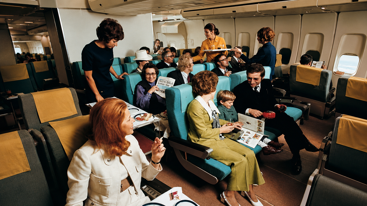 Passengers on an airplane enjoy meals and converse with each other. Flight attendants serve food. People are reading menus, smiling, and dressed in vintage 1970s clothing. The cabin appears spacious and well-lit.