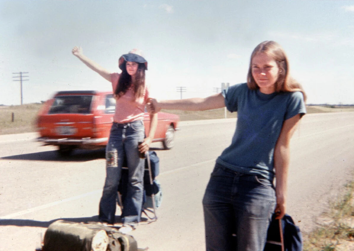 Two young women hitchhiking on a rural road, one closer to the camera in a blue shirt, the other in a hat and pink shirt, with a red car passing by and suitcases on the ground.