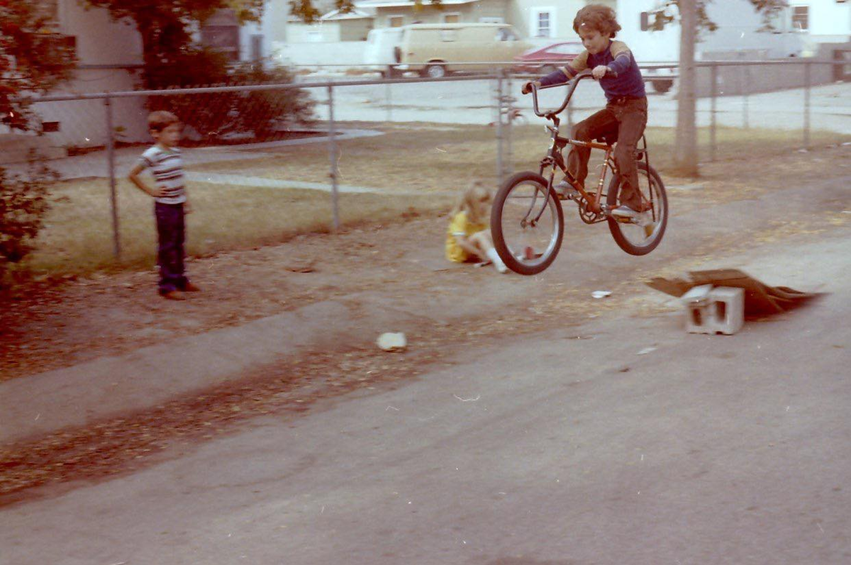A child on a bicycle jumps over a makeshift ramp on a street, while another child stands nearby and a third child sits on the grass, watching. Houses and cars are visible in the background.