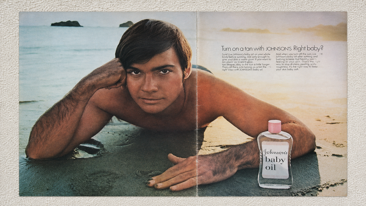 A man lies on sandy beach at sunset, propped on one elbow beside a bottle of Johnson’s baby oil. The text promotes using the oil for tanning. The sea and rocks are visible in the background.