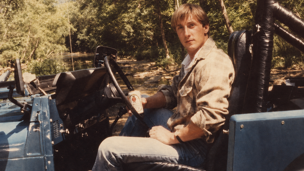 A man in casual clothes sits in a blue off-road vehicle by a shallow creek in a wooded area, holding a can and looking at the camera.