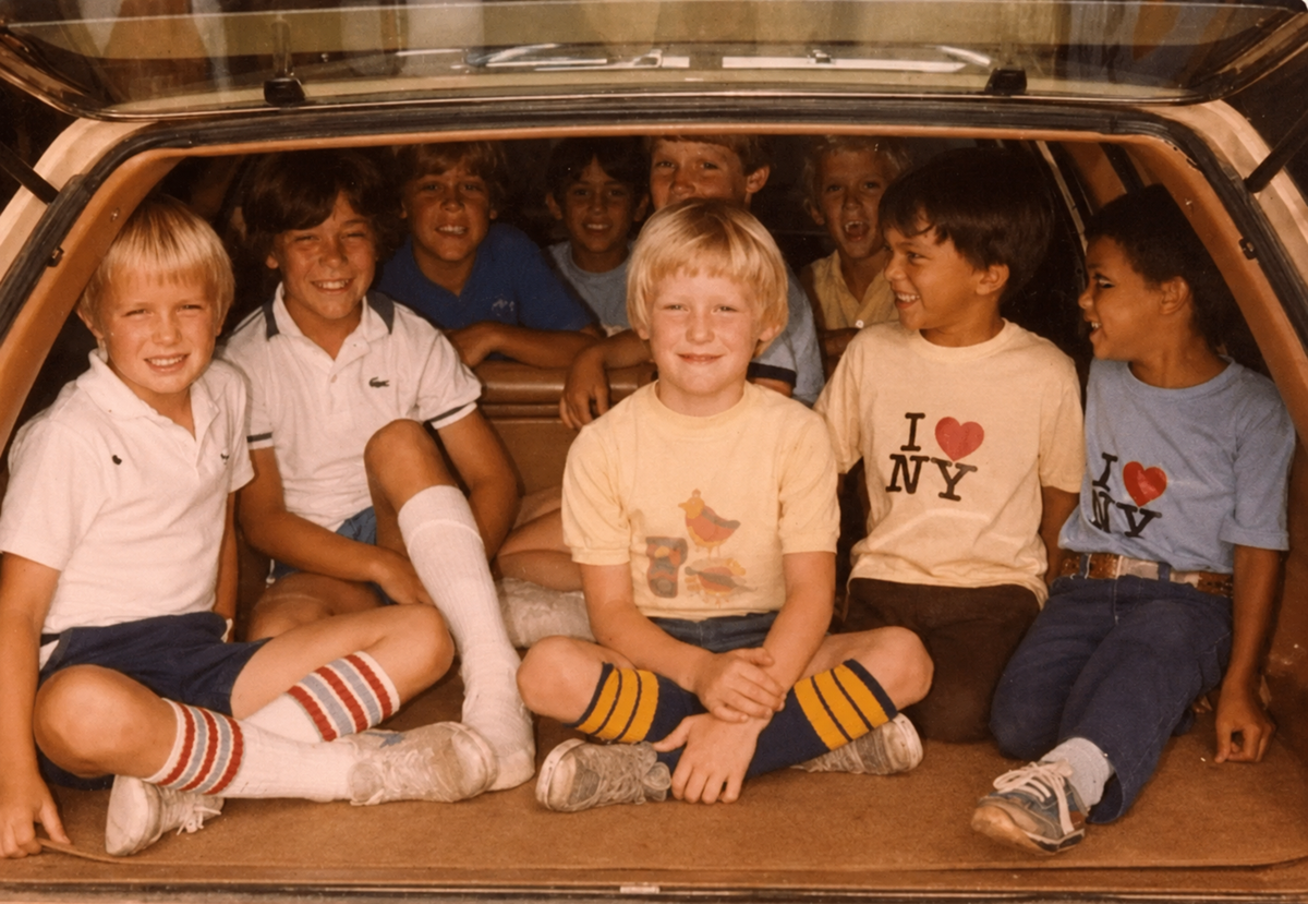 A group of smiling kids sit together in the open trunk of a car, wearing colorful shirts and striped socks. The boys are relaxed and cheerful, some wearing "I ♥ NY" shirts, enjoying a fun moment together.