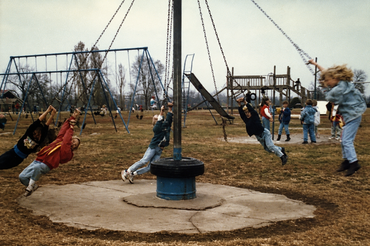 Four children spin on a playground merry-go-round swing, holding onto chains. Other kids play on swings and climbing equipment in the background. The scene is outdoors on a cloudy day.