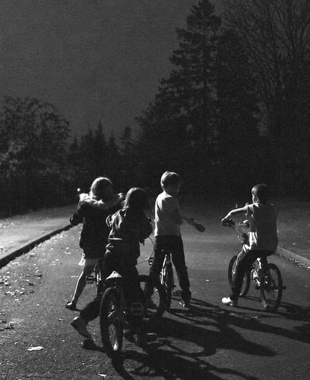 Four children ride bicycles together on a quiet street at night, surrounded by silhouettes of trees. The scene is lit by a soft light, creating long shadows on the ground.
