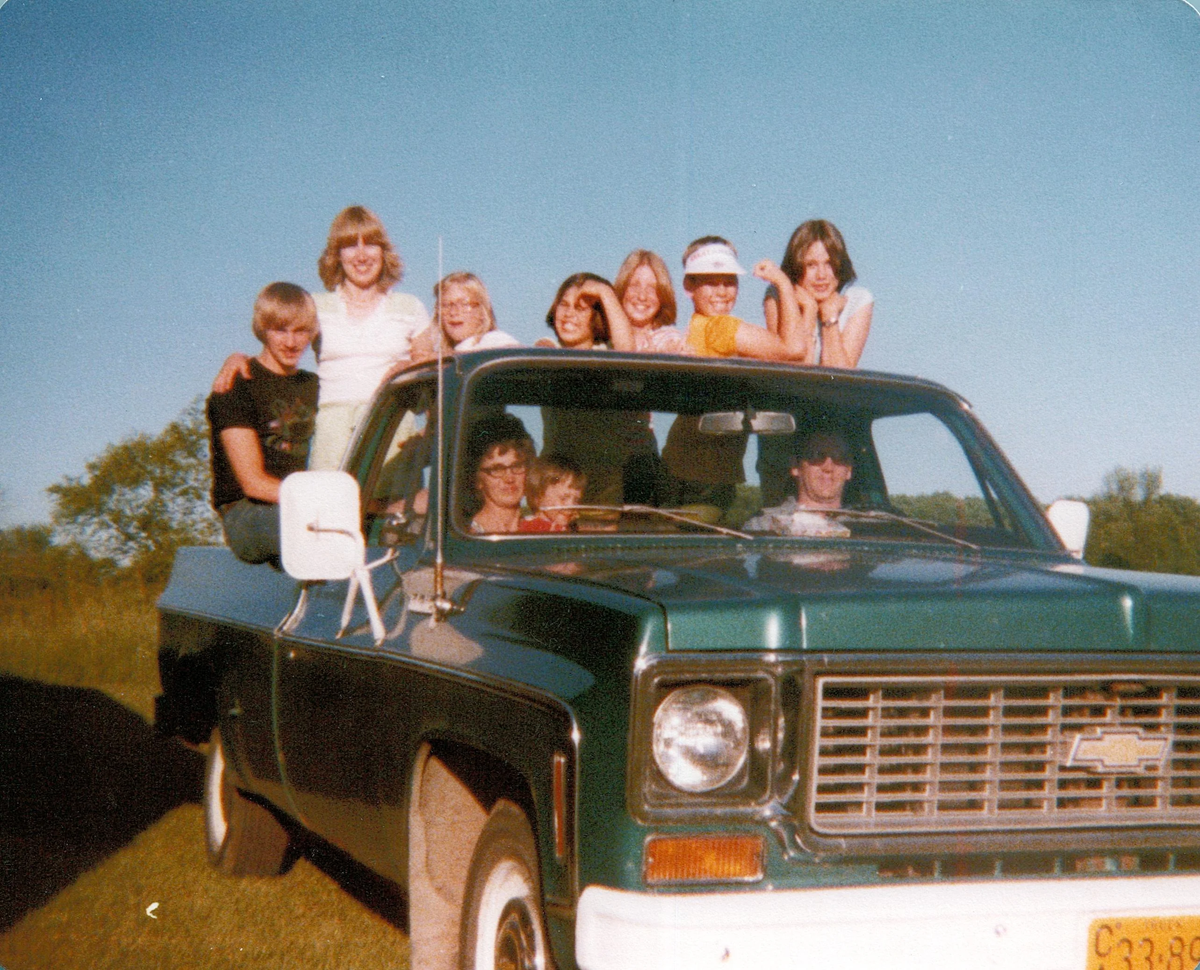 A group of smiling kids sits and stands on a green vintage Chevrolet truck parked in a grassy field under a clear blue sky.