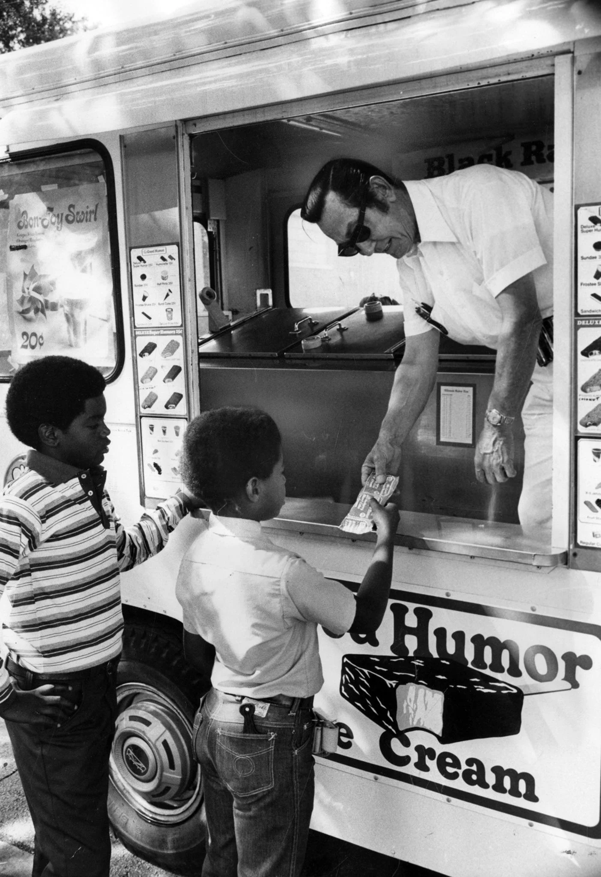 Two boys buy ice cream from a vendor in an ice cream truck. One boy hands money to the vendor, who is wearing sunglasses. Menus and ice cream posters are visible on the side of the truck.