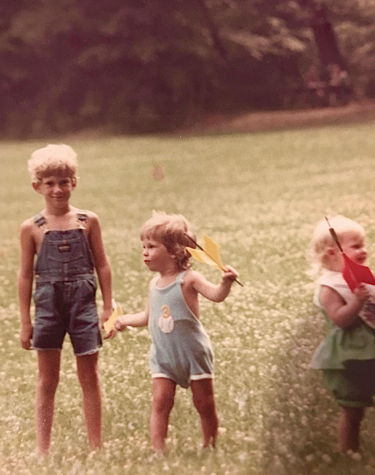 Three young children stand in a grassy field holding colorful toy planes. The boy on the left wears denim overalls, while the two girls wear light-colored outfits. Trees and greenery are visible in the background.