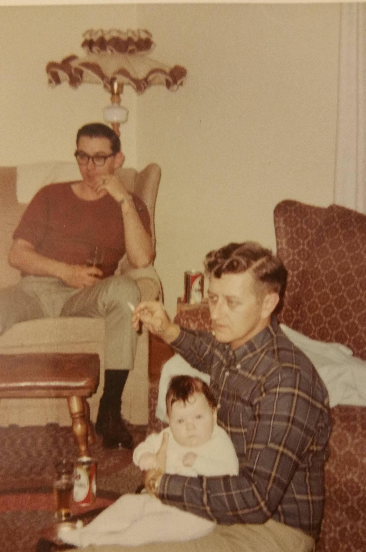 Two men sit in a living room; one holds a baby on his lap. Beers and glasses are on the table beside them. The background includes a patterned couch, lamp, and neutral-colored walls.