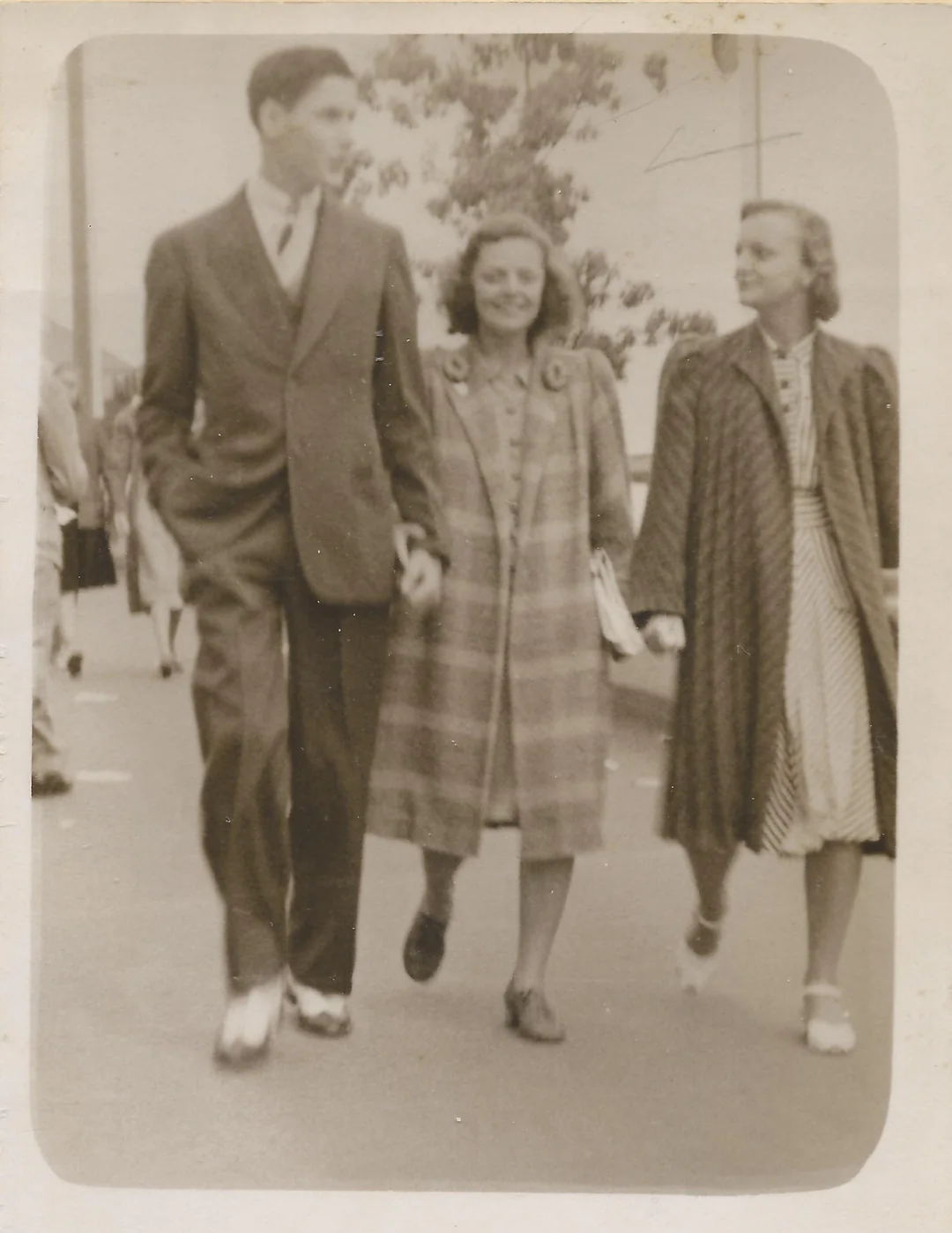 Three people walk down a street together, smiling. The man on the left wears a suit, while the two women beside him wear long coats. The photo is black and white and appears to be from the mid-20th century.