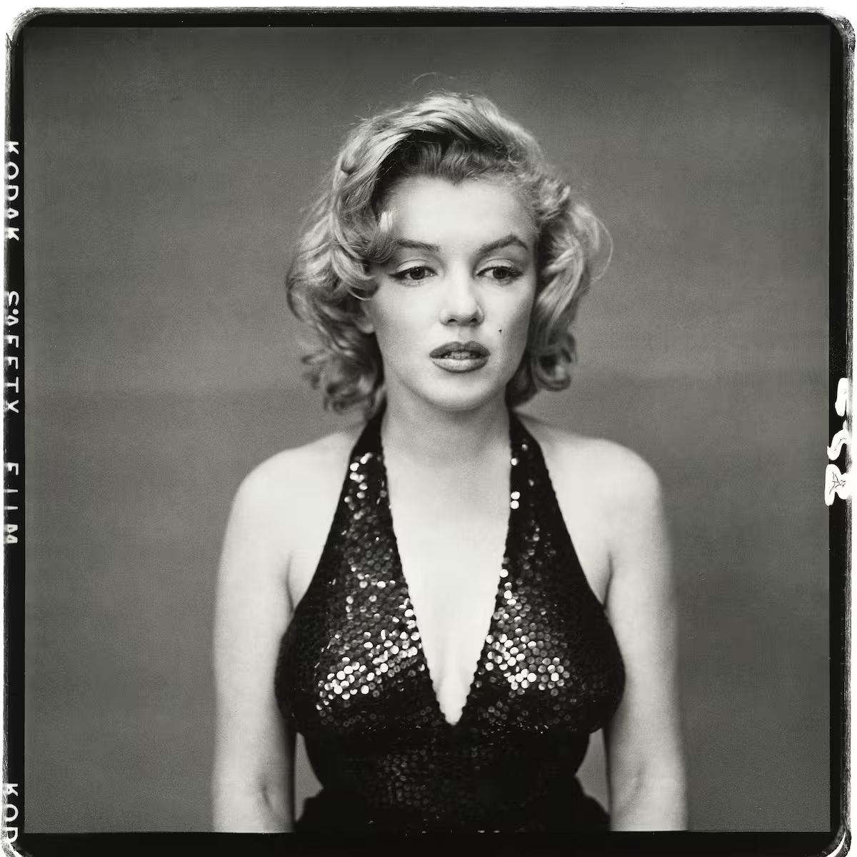 A woman with short, wavy blonde hair wears a sequined halter dress and looks slightly downward. The photo is black and white with a plain background, and film edges are visible.