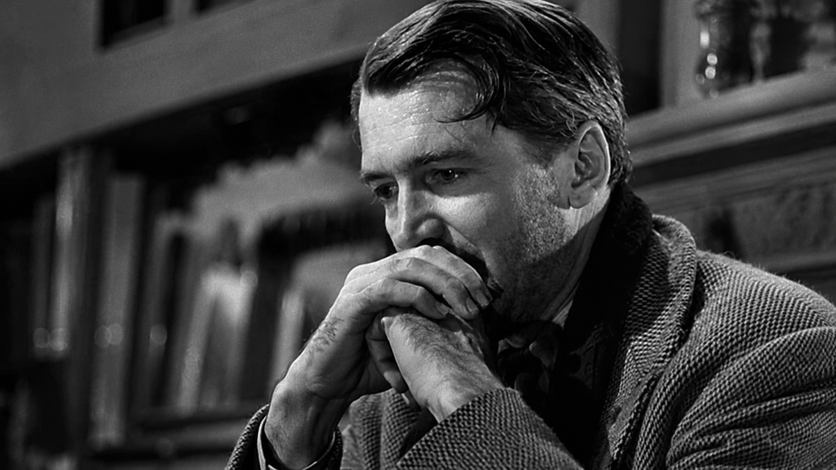 A man in a suit sits with his hands clasped near his mouth, looking thoughtful and concerned. The image is in black and white.