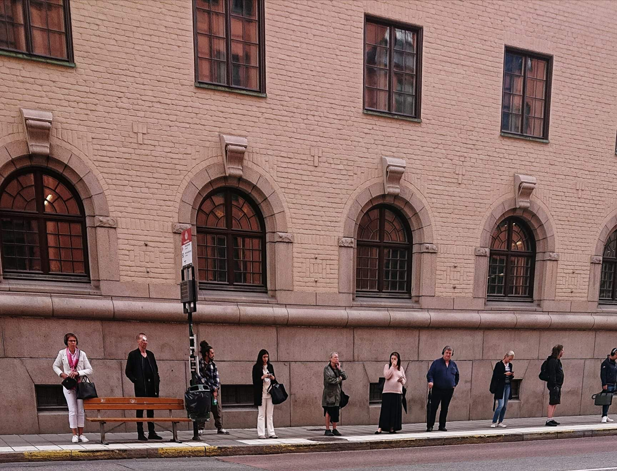 People stand spaced apart along a city sidewalk in front of a tan brick building with arched windows, appearing to wait at a bus stop. A wooden bench and a bus stop sign are visible.