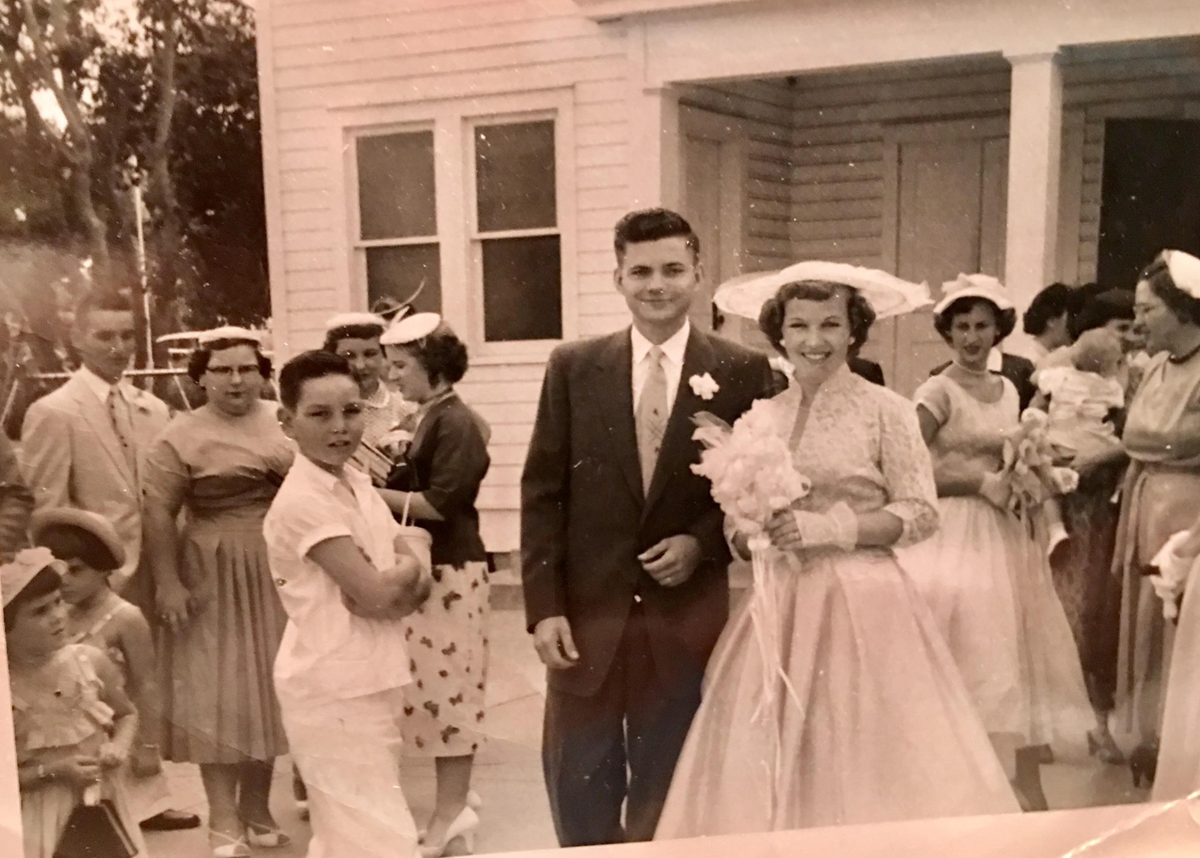 A vintage wedding photo shows a bride and groom smiling outside a house, surrounded by guests in 1950s attire, including women in dresses and hats, and children. The scene is casual and joyful.