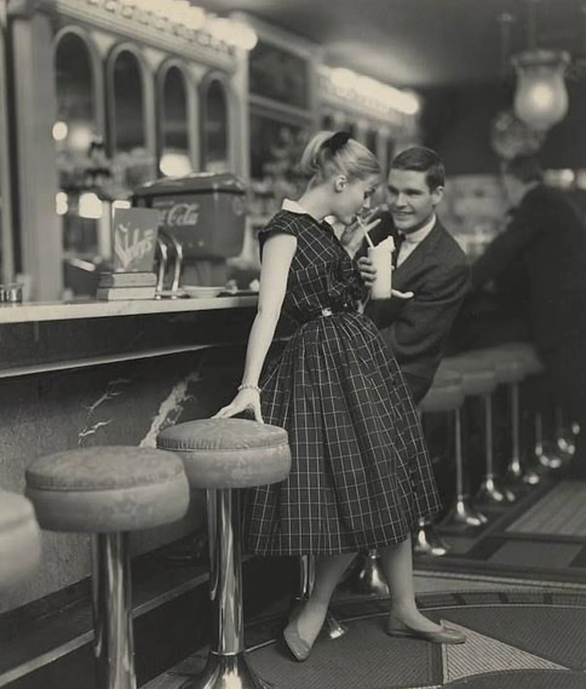 A young woman in a checkered dress shares a milkshake with a man in a suit at a retro diner counter, with vintage stools and decor visible.