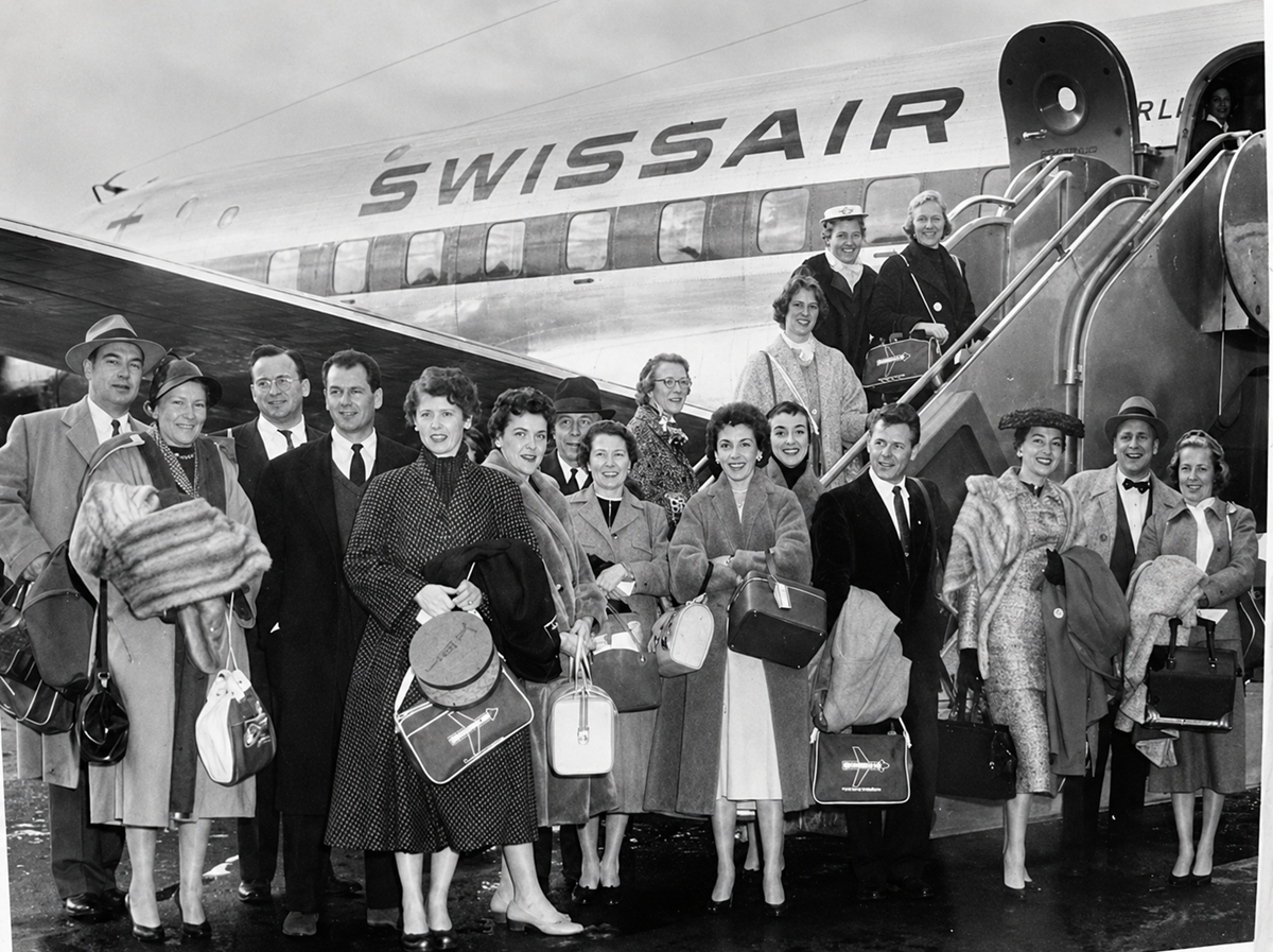 A group of people in 1950s-style clothing stands smiling in front of a Swissair airplane, holding bags and luggage, with some on the plane’s stairs and others on the tarmac.