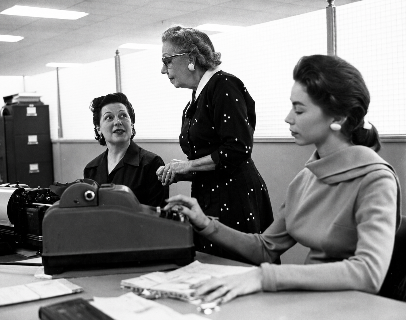 Three women in an office, two seated at typewriters and one standing between them, all wearing vintage 1950s clothing. Papers and office supplies are visible on the desks.